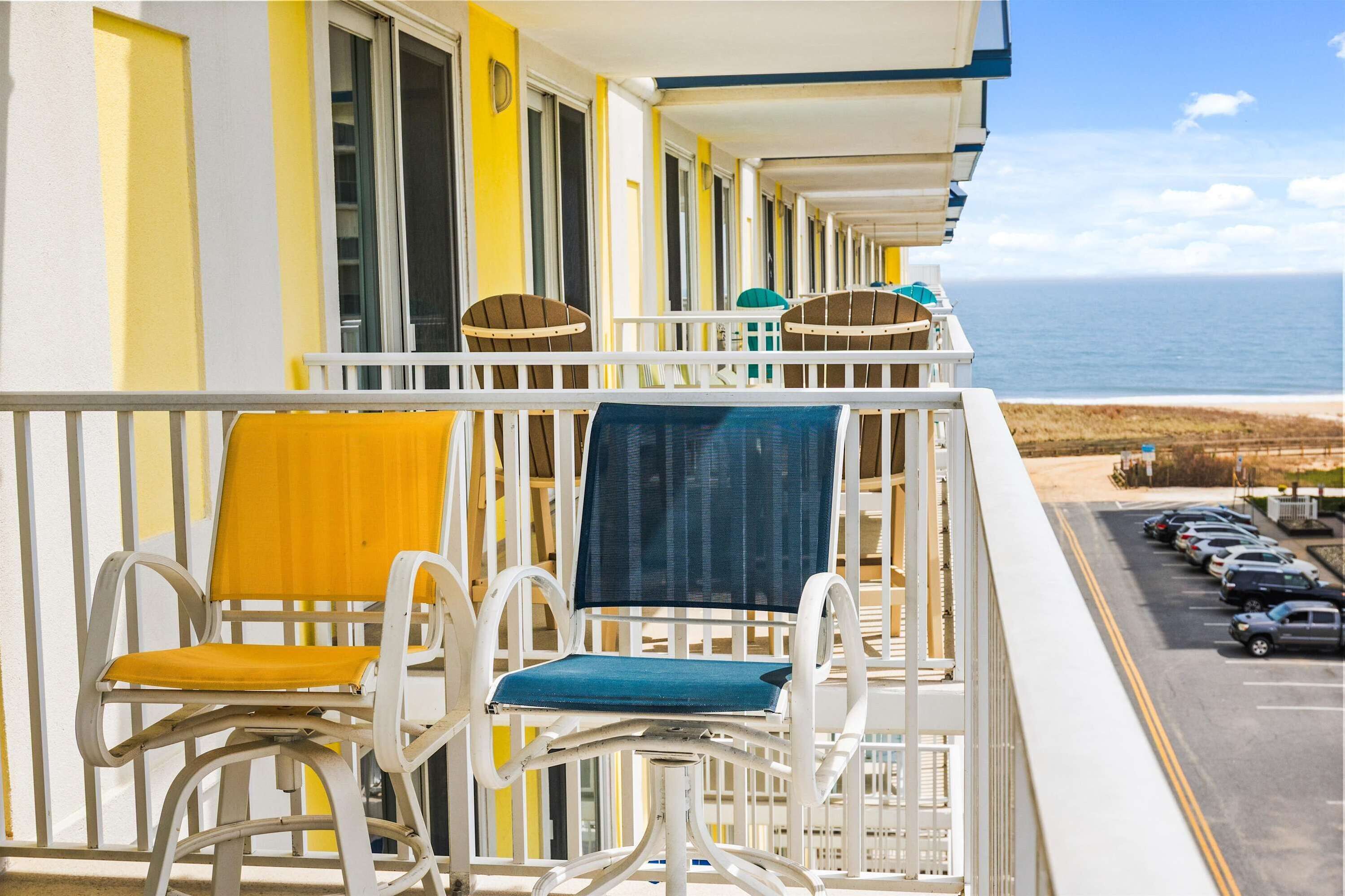 A balcony with two chairs, one with a yellow cushion and one with a blue cushion, overlooking a parking lot and the ocean.