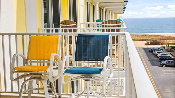 Un balcon avec deux chaises, dont une avec un coussin jaune et l’autre avec un coussin bleu, donnant sur un parking et l’océan.
