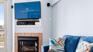 Un salon doté d’une cheminée, d’un canapé bleu agrémenté de coussins à motifs et d’un téléviseur à écran plat fixé au-dessus de la cheminée, qui diffuse une publicité pour « OCEANS EDGE VACATIONS ».