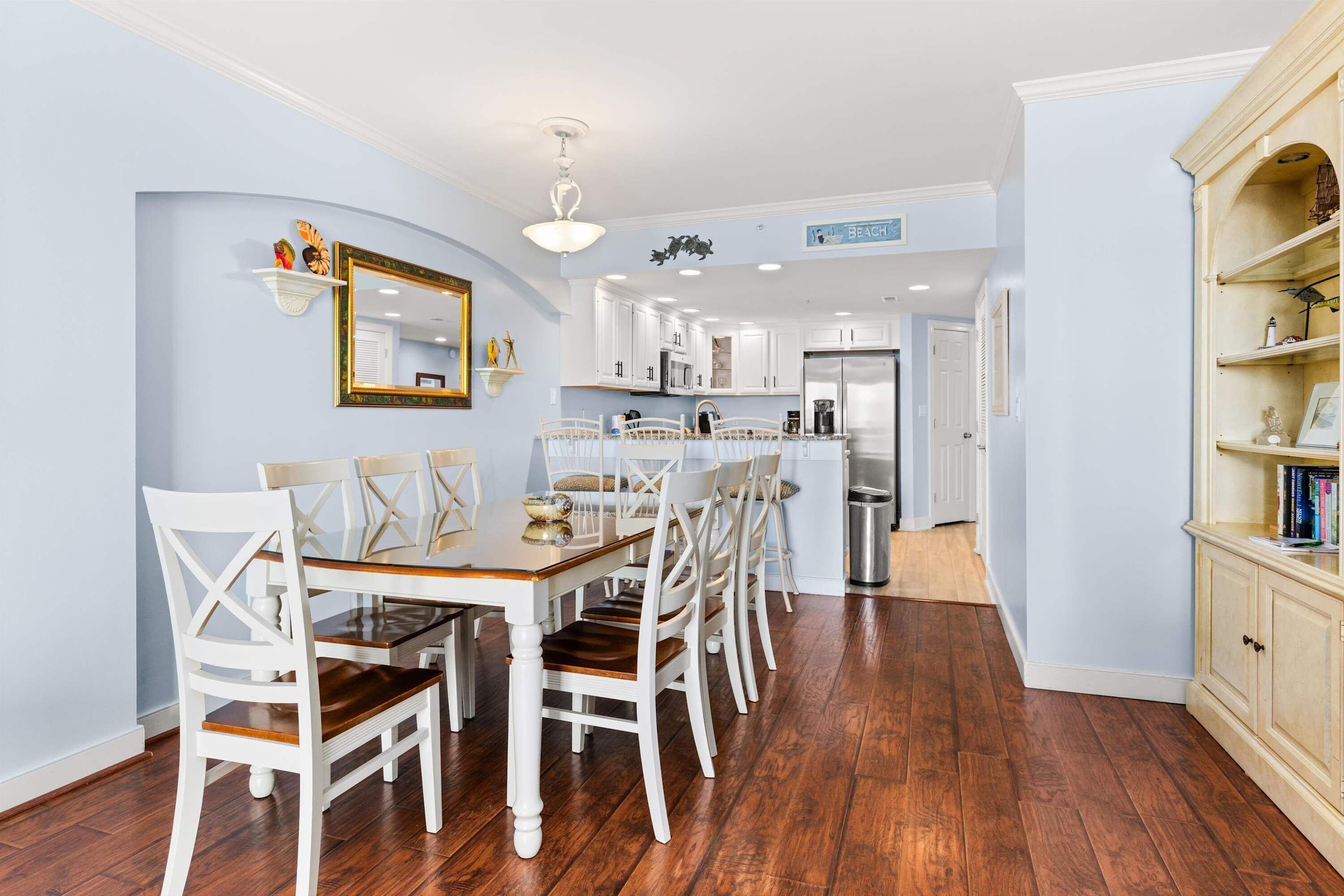 A dining area with a wooden table and white chairs, a kitchen with white cabinets, and a bookshelf with books.