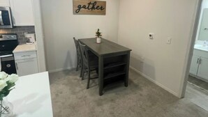 Une cuisine compacte dotée d’appareils électroménagers modernes, un coin repas avec une table et des chaises en bois foncé, et un panneau décoratif sur le mur avec l’inscription « rassemblement ».