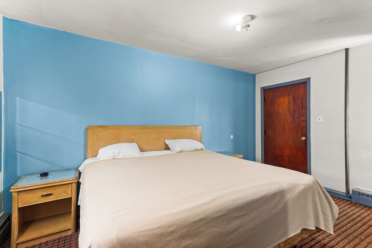A hotel room with a bed, a nightstand, a wooden door, and a light fixture on the ceiling.