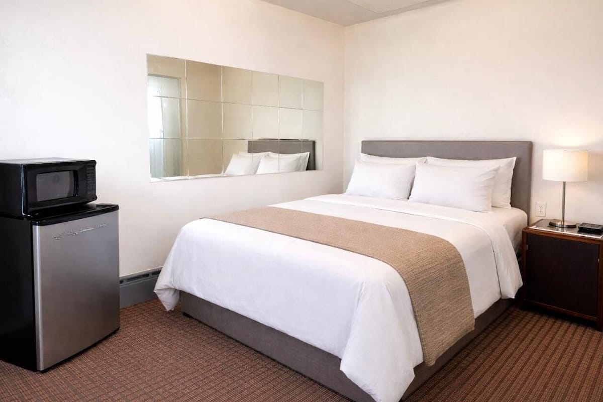 A hotel room with a bed, a small refrigerator, a bedside lamp, and a mirror on the wall.