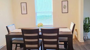 Un comedor con una mesa y cuatro sillas, una ventana con persianas y dos cuadros en la pared.