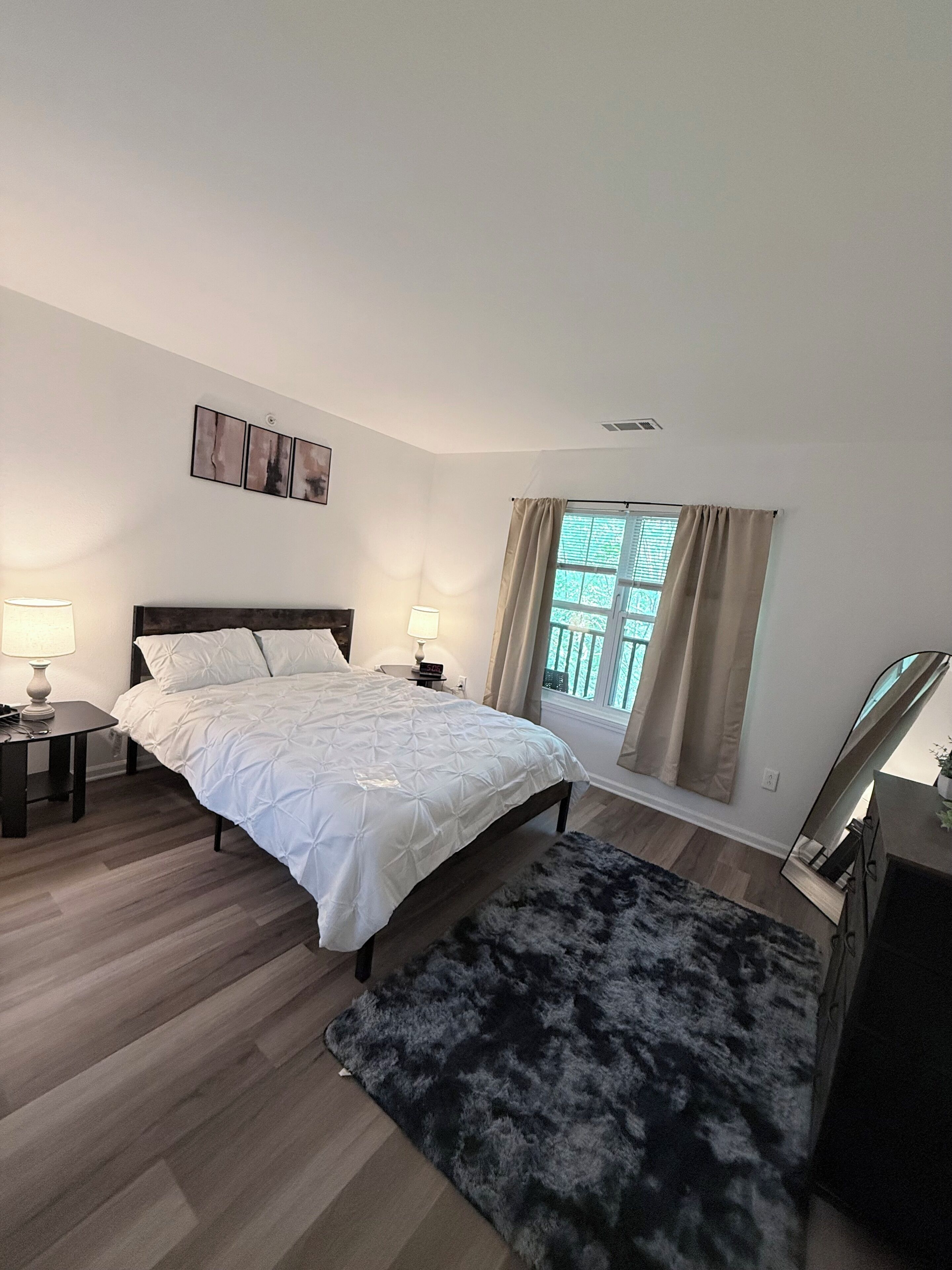 A bedroom with a bed, two bedside tables with lamps, a window with curtains, and a mirror.