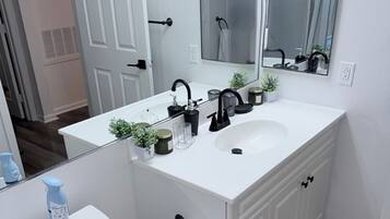 A bathroom with a white sink, black faucet, and a mirror above it. There is a toilet with a white lid, a trash can, and a green rug on the floor.
