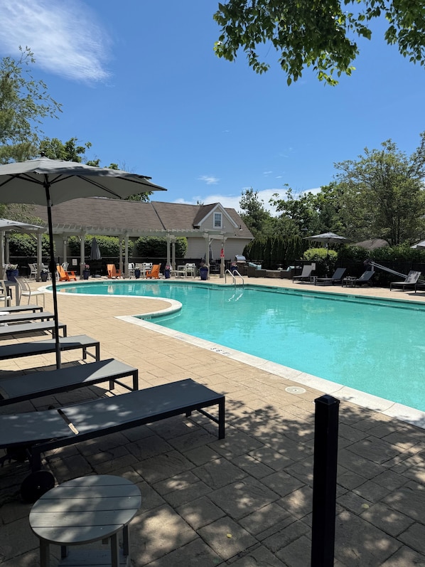Una piscina con sillas de descanso y sombrillas, un patio techado y una casa al fondo.