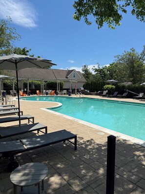 Une piscine avec des chaises longues et des parasols, un patio couvert et une maison en arriĂšre-plan.