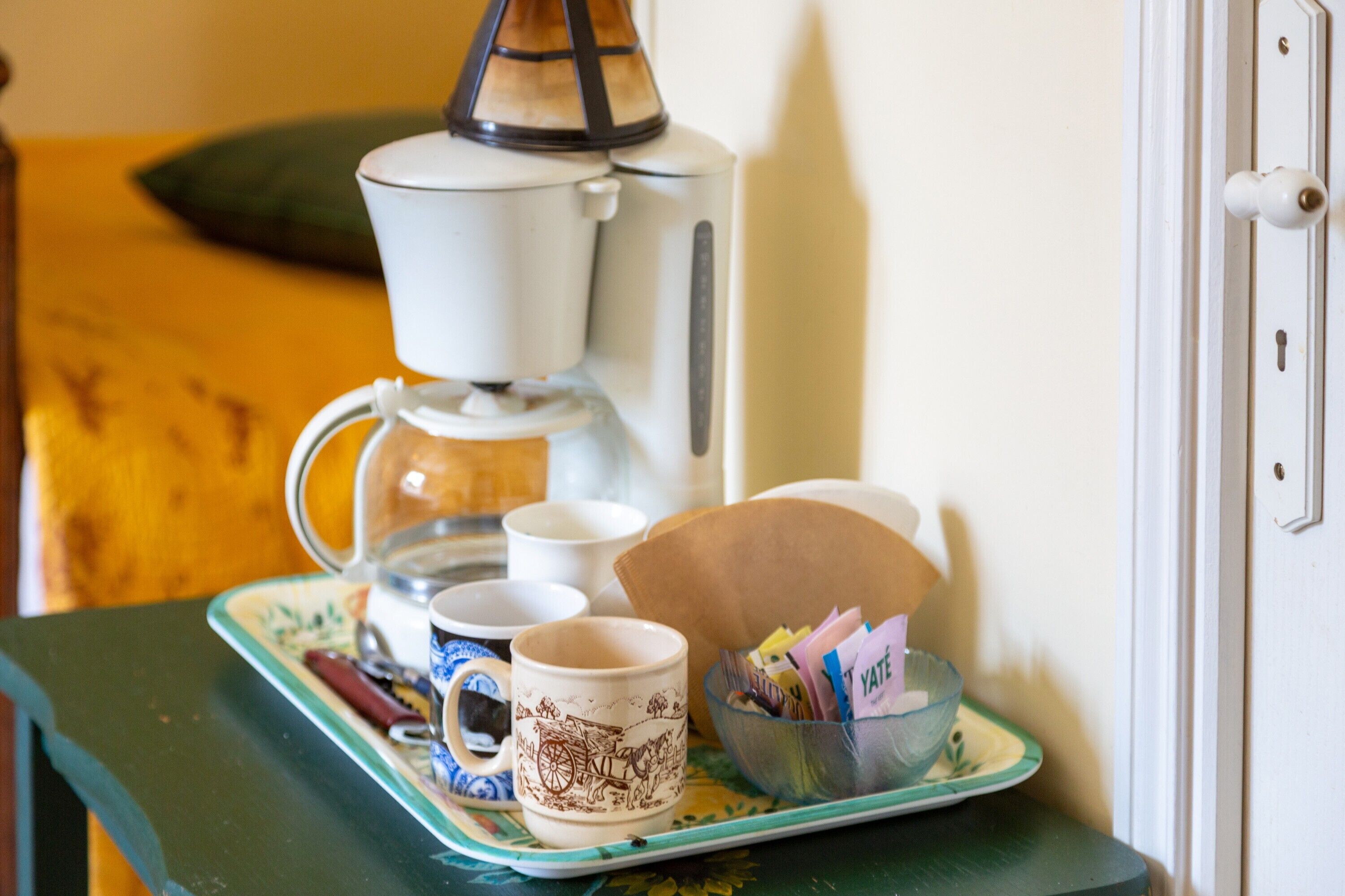 Una cafetera, tazas, una bandeja y un bol con correspondencia sobre una mesa.