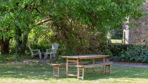 Una mesa de picnic de madera con bancos en un área verde con árboles y un muro de piedra al fondo.