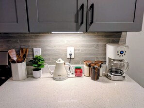 A kitchen with grey cabinets, a white countertop, a coffee maker, a kettle, a plant, and spice jars.