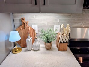 A kitchen counter with a lamp, two wine bottles, a potted plant, a wooden cutting board, and a knife block.