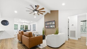 Ein modernes Wohnzimmer mit Holzboden, Deckenventilator, flachbildfernseher an einer Holzvertäfelung und einer Sitzecke mit braunem Ledersofa und weißem Sessel.