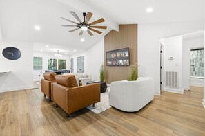 A modern living room with a wooden floor, a ceiling fan, a flat-screen TV mounted on a wooden panel, and a seating area with a brown leather sofa and a white armchair. - The Hidden Gem in North Myrtle Beach (North Myrtle Beach)