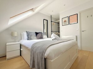 A modern bedroom with a sloped ceiling, a bed with a grey blanket, a bedside table with a lamp, and a wardrobe. - Camden Town Apartment by PrimeCityHosts (Greater London)