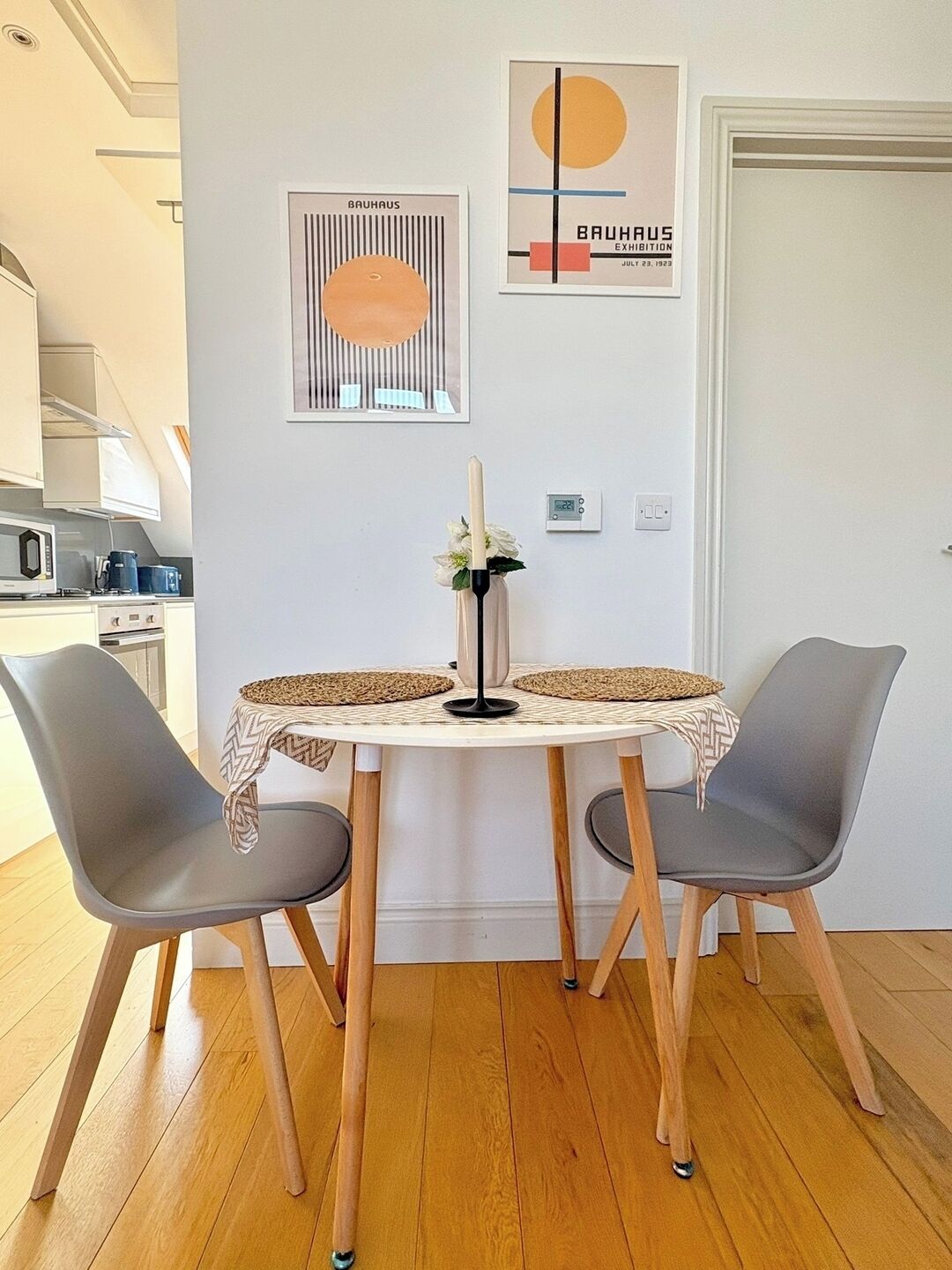 A dining area with a round table, two chairs, a candle centerpiece, and two framed artworks on the wall.