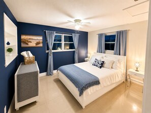 Ein Schlafzimmer mit einem Bett, einem Nachttisch, einem an der Wand befestigten Regal, einem Deckenventilator und einem Fenster mit Vorhängen.