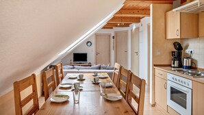 Ein Essbereich mit Holztisch, gedeckt für vier Personen, eine Küche mit Herd und Schränken sowie ein Wohnbereich mit Sofa und Fernseher.
