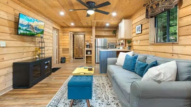 L’intérieur d’un chalet en bois comprend un salon, un canapé, un pouf, un ventilateur de plafond, un téléviseur, un coin cuisine et une porte donnant sur une autre pièce.