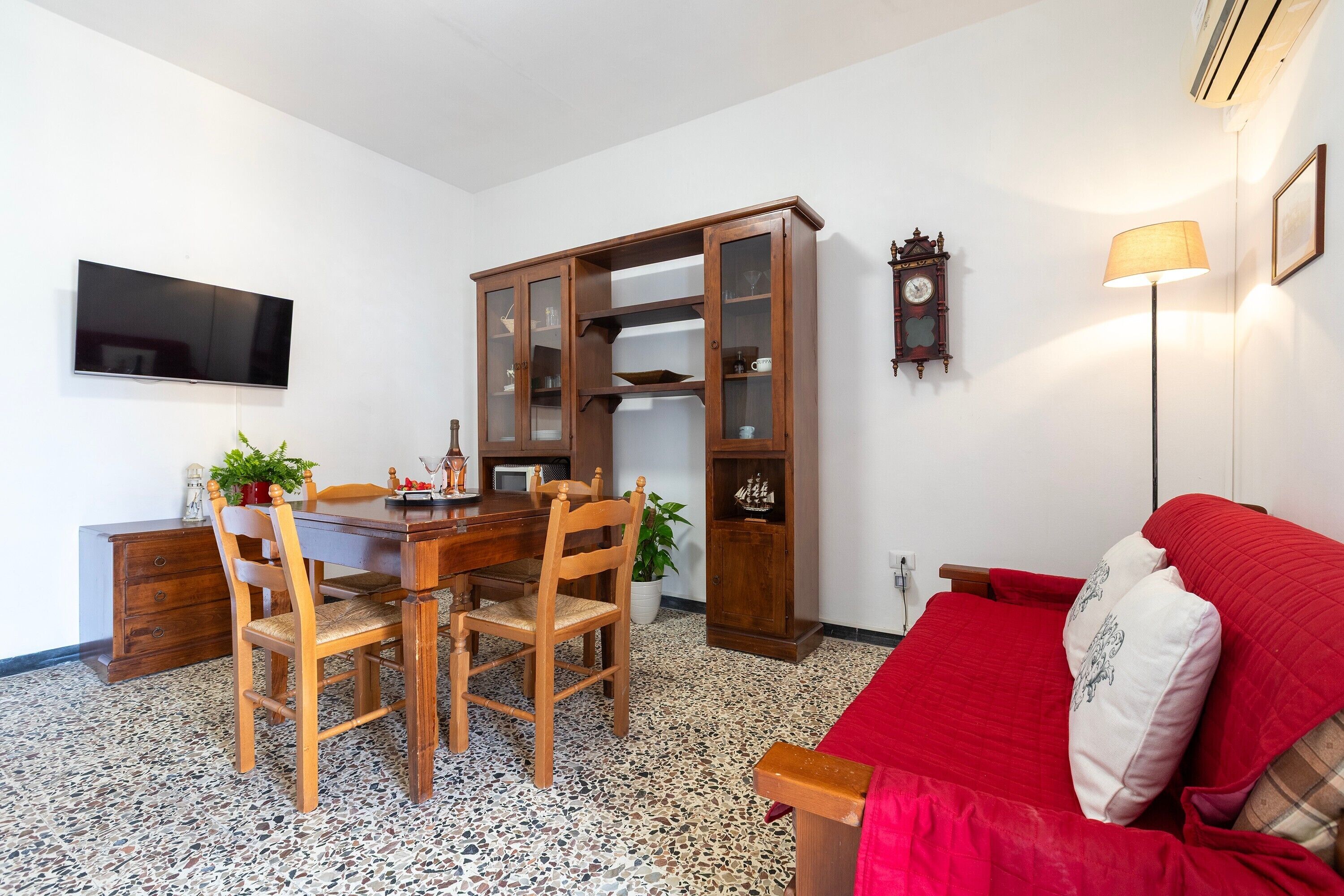 A room with a dining table, chairs, a red sofa, a TV, a cabinet, a clock, and a lamp.