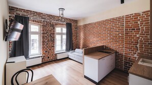 A room with brick walls, a white sofa, a small kitchen area, and a window with curtains. - Apartament Ceglane Studio (Toruń)