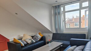 A modern living room with a black leather sofa, a wooden coffee table, and a view of buildings through a window. - Spacious Apartment Sleeps 7 near central London (Greater London)