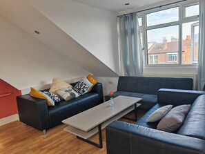 A modern living room with a black leather sofa, a wooden coffee table, and a view of buildings through a window. - Spacious Apartment Sleeps 7 near central London (Greater London)