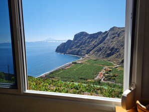 A coastal view through a window, including a beach, mountains, and a distant mountain peak.