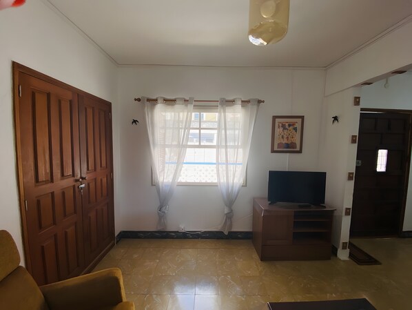 A room with a wooden door, a television on a stand, a chair, a window with curtains, and a ceiling light fixture.
