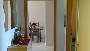Een smalle gang die uitkomt op een eetruimte met een tafel en stoelen, een kleine plant op een bijzettafeltje, en een deur aan de rechterkant.