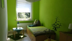 Een kamer met een bed, een klein tafeltje, een radiator, een raam met uitzicht op bomen en een plant.
