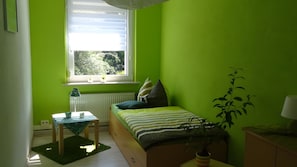 Ein Zimmer mit einem Bett, einem kleinen Tisch, einem Heizkörper, einem Fenster mit Blick auf Bäume und einer Pflanze.
