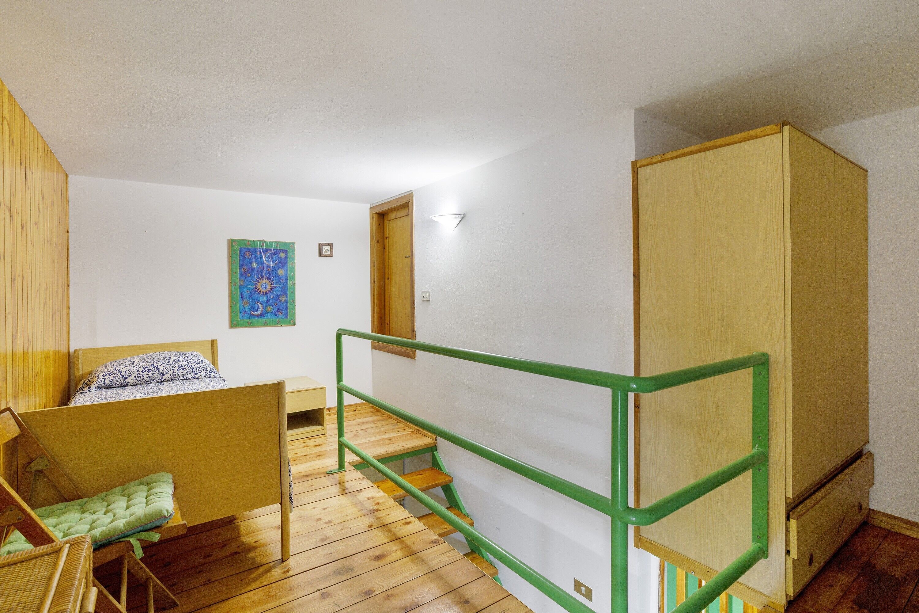 Une chambre avec un lit superposé, une rambarde en métal vert, une armoire en bois et un petit tableau accroché au mur.