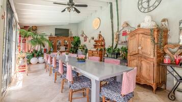 Un coin repas avec une longue table, des chaises à carreaux, un ventilateur de plafond et un cactus accroché au mur.
