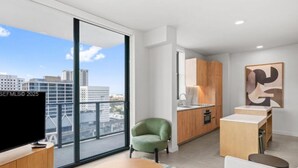 Um apartamento moderno com uma janela ampla, televisão de tela plana, uma cadeira verde, uma mesa de jantar de madeira e uma área de cozinha.