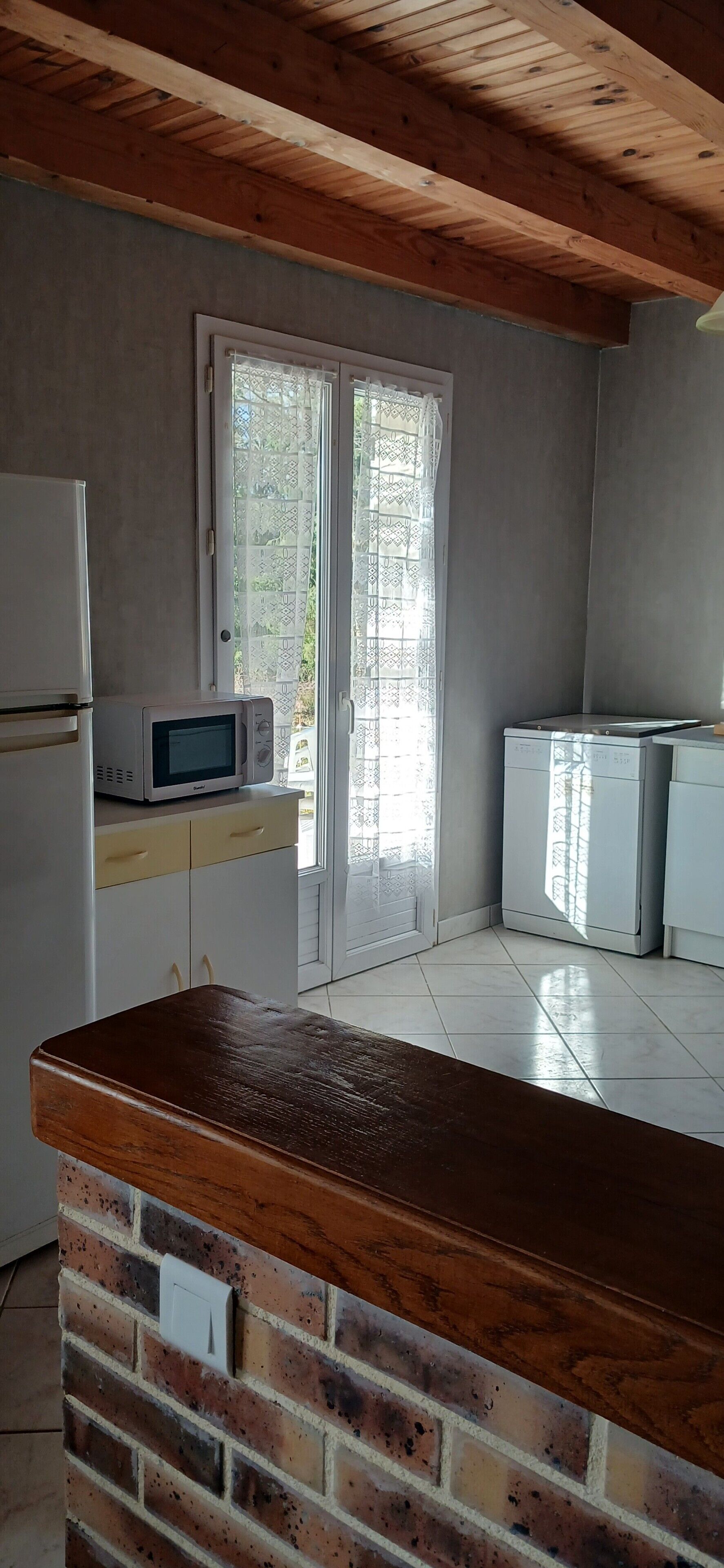 A kitchen with a microwave, refrigerator, and cabinets. There is a wooden ceiling and a brick wall.