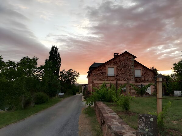 Une maison en pierre avec une allée pavée, entourée de verdure et sous un ciel nuageux.