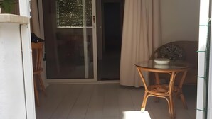 Una habitación con puerta de vidrio corredera que da a un balcón, una mesa redonda con sillas y un estante pequeño fijado a la pared.