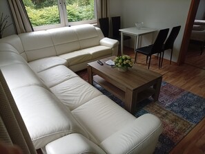 A living room with a white leather sofa, a wooden coffee table with a vase of flowers, and a dining area with a small table and chairs.