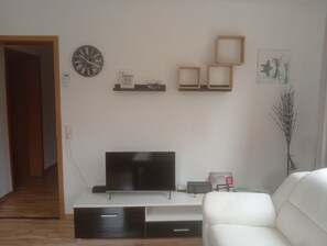 Un salon avec un téléviseur à écran plat posé sur un meuble, un canapé blanc, une porte en bois et une horloge murale.