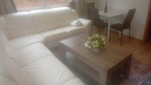 En stue med en hvid sofa, et træsofabord med en vase blomster, et spisebord med stole, og udsigt gennem et vindue.