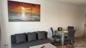 A modern living room with a grey sofa, a coffee table, a dining area with a small table and chairs, and a large painting of a sunset over the ocean. - Studio Apartment 'Luv Lee Duhnen 02' with Private Terrace, Shared Garden and Wi-Fi (Cuxhaven)