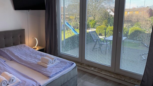 A hotel room with a bed, a TV, and large windows with blinds. - Studio Apartment 'Luv Lee Duhnen 02' with Private Terrace, Shared Garden and Wi-Fi (Cuxhaven)