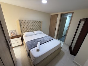 A bedroom with a tufted headboard, a bed with white linens, a grey blanket, and a rolled towel. There is a bedside table with a framed picture, a wooden wardrobe, and a door leading to another room.