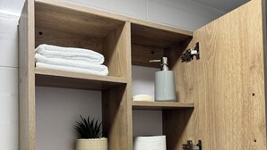 A wooden cabinet with shelves containing a towel, a bottle, a plant, and toilet paper.