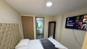 A hotel room with a tufted headboard, a bed with white and dark grey bedding, a bathroom visible through an open door, and a TV mounted on the wall displaying movie posters.