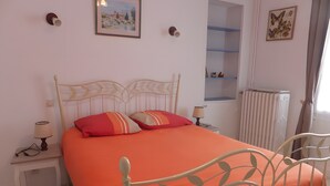 Una cama con colcha naranja, cabecera de metal blanco, dos lámparas de noche, una mesita con lámpara, y un cuadro enmarcado en la pared.
