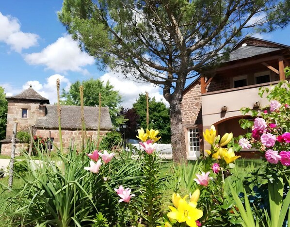 A stone building with a thatched roof, surrounded by a garden with various flowers and a large tree.