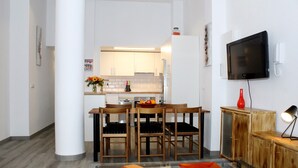 Eine moderne Küche mit weißen Schränken, einem Esstisch mit Stühlen, einem an der Wand befestigten Fernseher und einer kleinen Kommode mit einer Lampe.