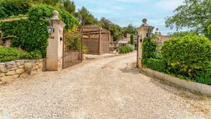 Une allée de gravier mène à une entrée fermée par un portail, agrémentée d’une pergola et d’un mur en pierre.